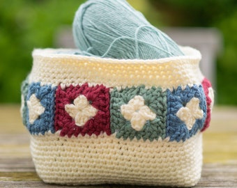 May include: A cream-colored crochet basket with a band of colorful squares. Each square features a white flower design. The basket holds a ball of light blue yarn, perfect for storing craft supplies.