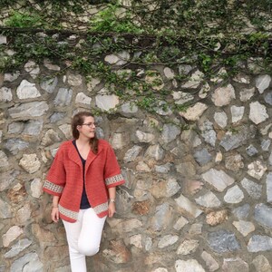 Peut inclure: Une femme portant un cardigan en crochet corail avec des bordures blanches, un pantalon blanc et des chaussures marrons. Elle se tient devant un mur de pierre recouvert de vignes vertes.