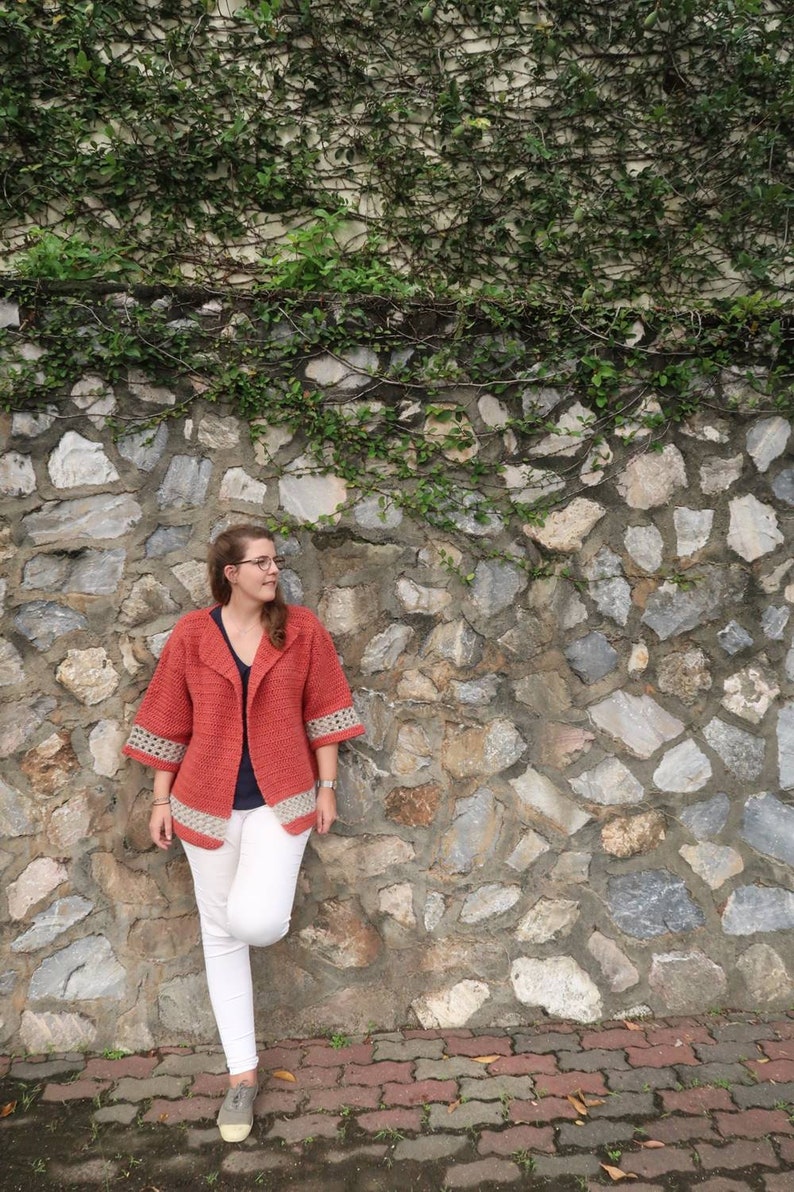 Peut inclure: Une femme portant un cardigan en crochet corail avec des bordures blanches, un pantalon blanc et des chaussures marrons. Elle se tient devant un mur de pierre recouvert de vignes vertes.