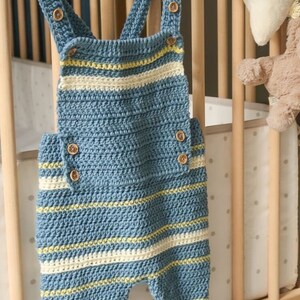 May include: A crochet baby romper with blue and white stripes and yellow accents. The romper has buttons on the straps and is hanging on a wooden hanger. The text "PRINTABLE PDF CROCHET PATTERN" is at the bottom of the image.