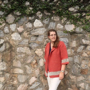 Peut inclure: Une femme portant un cardigan en crochet corail avec une bordure en crochet blanc. Elle se tient devant un mur en pierre avec des vignes qui poussent dessus.