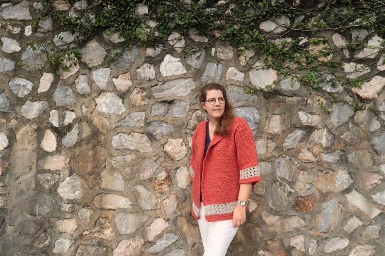 Peut inclure: Une femme portant un cardigan en crochet corail avec une bordure en crochet blanc. Elle se tient devant un mur en pierre avec des vignes qui poussent dessus.