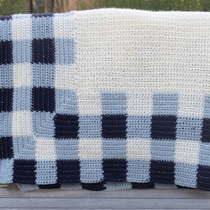 May include: A handmade crochet blanket with a checkerboard pattern in shades of blue and white. The blanket is draped over a wooden surface, showcasing its texture and design. The top portion is solid white.