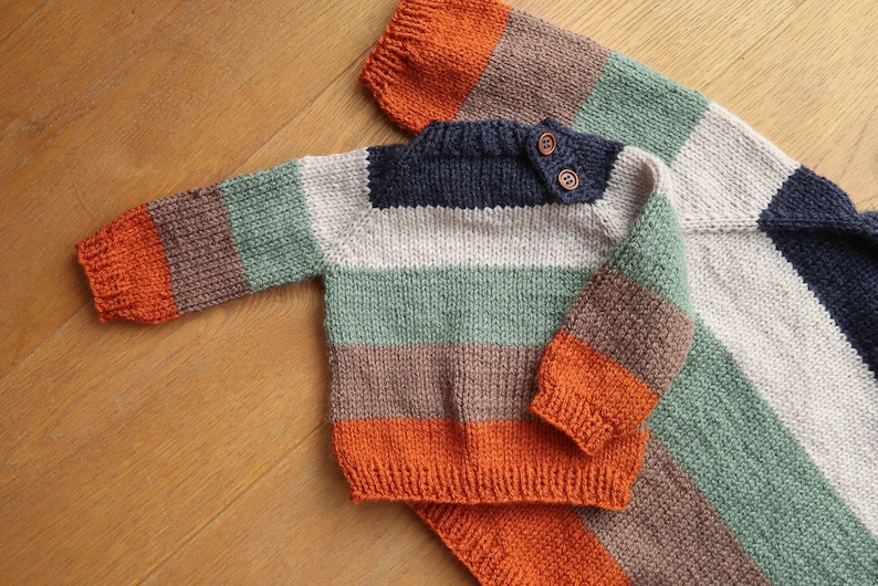 Op de afbeelding: Een gestreepte gebreide trui in tinten oranje, bruin, groen, wit en donkerblauw. De trui heeft een ronde hals en lange mouwen.