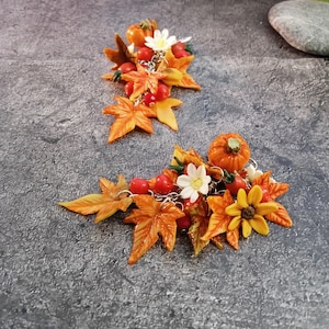 Op de afbeelding: Twee haarclips met herfstthema. Elke clip heeft oranje esdoornbladeren, kleine pompoenen, rode bessen en witte en gele bloemen. De clips zijn verbonden door zilveren kettingen.