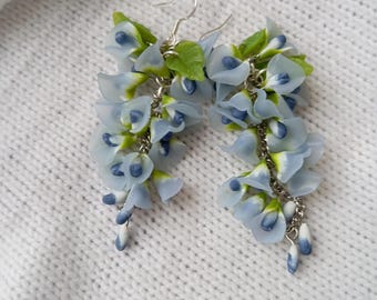 May include: A pair of dangle earrings featuring light blue wisteria flower clusters with green leaves. The earrings have silver-colored hooks and chains, and are set against a white, textured background.
