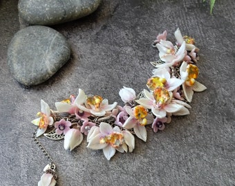 May include: A floral necklace featuring white and pink orchid-like flowers with yellow and orange centers. The necklace has a silver chain and a dangling flower pendant. The design includes small purple flowers and silver filigree accents.