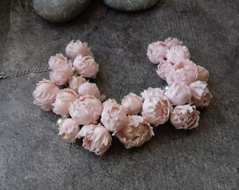 May include: A necklace featuring a series of light pink, fabric flower blossoms. The blossoms are arranged in a curved shape and interspersed with small, white beads. The necklace is set against a gray background.