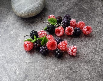 May include: Handmade earrings featuring detailed miniature red raspberries and blackberries with green leaves. The berries are clustered together, creating a realistic and colorful design. The earrings are on a gray surface.