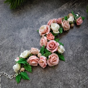 Pode incluir: Uma pulseira floral com rosas rosa e brancas e folhas verdes. A pulseira apresenta rosas em diferentes estágios de floração. Possui uma corrente prateada. O design é elegante e natural.