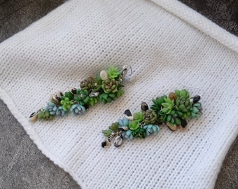 Puede incluir: Un par de pendientes hechos a mano con plantas suculentas en miniatura en tonos verdes, azules y beige. Los pendientes tienen ganchos plateados y están colocados sobre una superficie de punto blanca. Miden unos 5 cm.