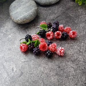 May include: A pair of handmade earrings with miniature raspberries and blackberries. The earrings display bright red and dark purple berries, with small green leaves. The jewellery is set against a textured grey background.