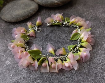 May include: A delicate bracelet featuring a floral design. The bracelet is adorned with light pink and green flower petals and leaves, connected by a silver chain. The flowers have a translucent quality, and the bracelet is set against a gray background.