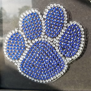 May include: A decorative, flower-shaped embellishment, densely covered with small, round, blue and clear rhinestones. The rhinestones are arranged in a pattern, with blue stones forming the petals and clear stones outlining the shape. The background is dark.