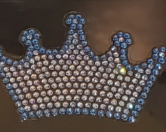 May include: A sparkling crown design, featuring a multitude of small, clear and blue rhinestones. The crown's outline and details are defined by the blue stones, creating a shimmering effect. The design is set against a dark background.