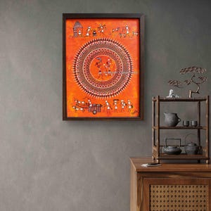 Pu&ograve; includere: Un dipinto rettangolare incorniciato con arte Warli su uno sfondo arancione vibrante. L'opera mostra un grande motivo a mandala circolare centrale circondato da piccole figure stilizzate bianche che raffigurano la vita quotidiana, animali e un carro.