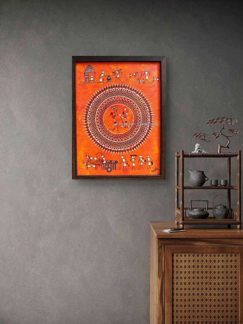 Pu&ograve; includere: Un dipinto rettangolare incorniciato con arte Warli su uno sfondo arancione vibrante. L'opera mostra un grande motivo a mandala circolare centrale circondato da piccole figure stilizzate bianche che raffigurano la vita quotidiana, animali e un carro.