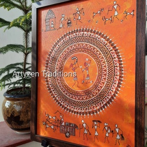 Pu&ograve; includere: Un dipinto incorniciato su sfondo arancione con figure stilizzate bianche in stile Warli. Il centro mostra un grande motivo a mandala circolare con due figure. Il testo Artyzen Traditions &egrave; stampato in bianco al centro.