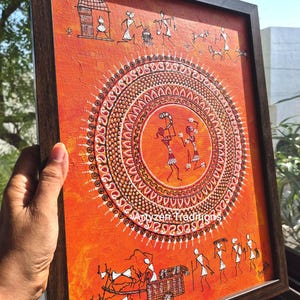 Pu&ograve; includere: Un dipinto rettangolare incorniciato su sfondo arancione con figure stilizzate bianche in stile Warli. L'opera mostra un motivo a mandala circolare centrale con due figure danzanti, circondato da scene di vita quotidiana e capanne.