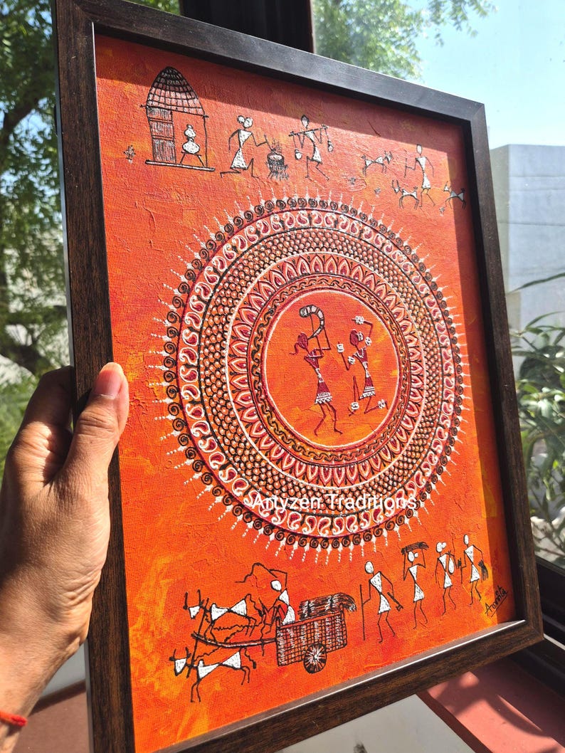 Pu&ograve; includere: Un dipinto rettangolare incorniciato su sfondo arancione con figure stilizzate bianche in stile Warli. L'opera mostra un motivo a mandala circolare centrale con due figure danzanti, circondato da scene di vita quotidiana e capanne.