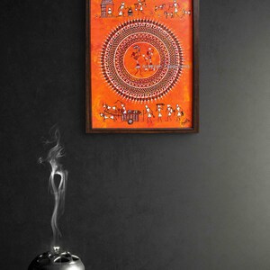 Pu&ograve; includere: Un dipinto rettangolare incorniciato che presenta figure stilizzate bianche in stile Warli su uno sfondo arancione vibrante. L'opera mostra un grande motivo mandala circolare centrale con figure danzanti, circondato da scene di vita quotidiana.