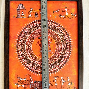 Pu&ograve; includere: Un dipinto rettangolare incorniciato su sfondo arancione con figure bianche in stile Warli, un motivo mandala circolare centrale e un righello in metallo che mostra un'altezza di circa 46 cm. L'opera raffigura la vita quotidiana del villaggio.