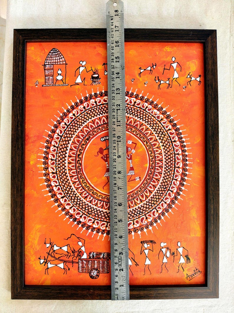 Pu&ograve; includere: Un dipinto rettangolare incorniciato su sfondo arancione con figure bianche in stile Warli, un motivo mandala circolare centrale e un righello in metallo che mostra un'altezza di circa 46 cm. L'opera raffigura la vita quotidiana del villaggio.