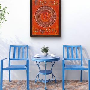 Pu&ograve; includere: Un dipinto rettangolare incorniciato con sfondo arancione che presenta intricate figure tribali bianche e un motivo centrale a mandala circolare. L'opera d'arte &egrave; esposta sopra due sedie in metallo blu brillante e un tavolo rotondo in metallo abbinato.