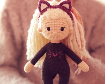 May include: A crocheted doll with blonde hair, wearing a purple cat headband and a black cat costume with a pink heart on the chest. The doll is holding a white tail.