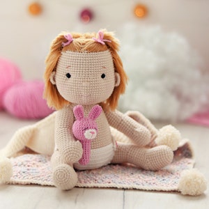 Peut inclure: Une poup&eacute;e en crochet aux cheveux bruns et aux n&oelig;uds roses est assise sur une couverture color&eacute;e. La poup&eacute;e tient un jouet en crochet rose en forme de lapin.