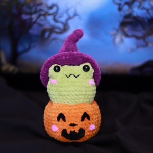 May include: A green crocheted frog wearing a purple witch hat sits on top of an orange crocheted pumpkin with a carved jack-o'-lantern face.
