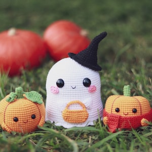 May include: A white crocheted ghost wearing a black witch hat holds a small orange crocheted pumpkin bucket. Two orange crocheted pumpkins with smiling faces are on either side of the ghost. All three are sitting on green grass.