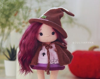 May include: A crocheted doll wearing a brown witch hat, a purple and pink dress, and brown cape. The doll has long, curly, burgundy hair and green shoes. The doll is standing on a white surface with a red pumpkin in the background.