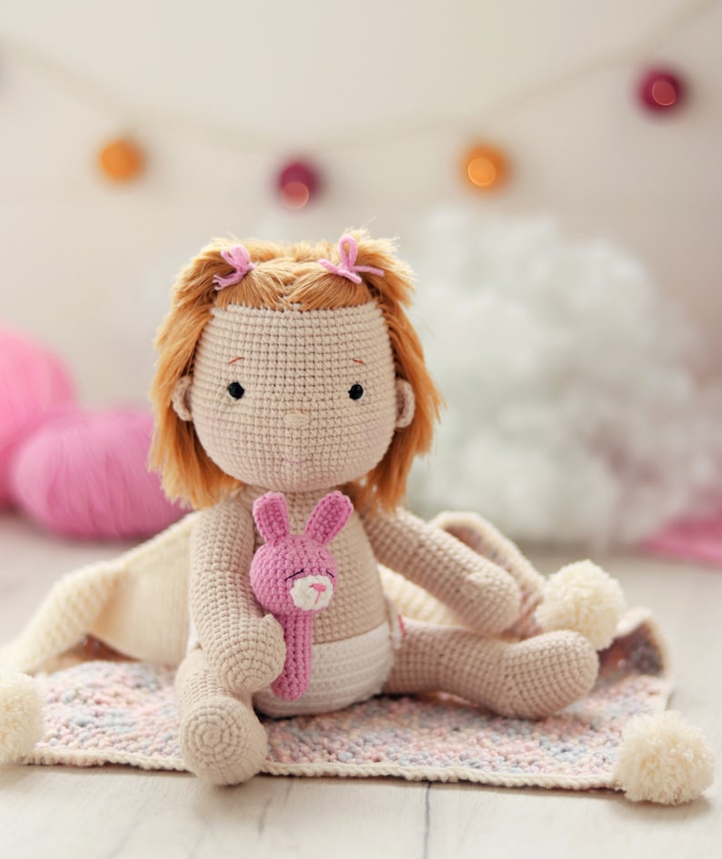 Peut inclure: Une poup&eacute;e en crochet aux cheveux bruns et aux n&oelig;uds roses est assise sur une couverture color&eacute;e. La poup&eacute;e tient un jouet en crochet rose en forme de lapin.