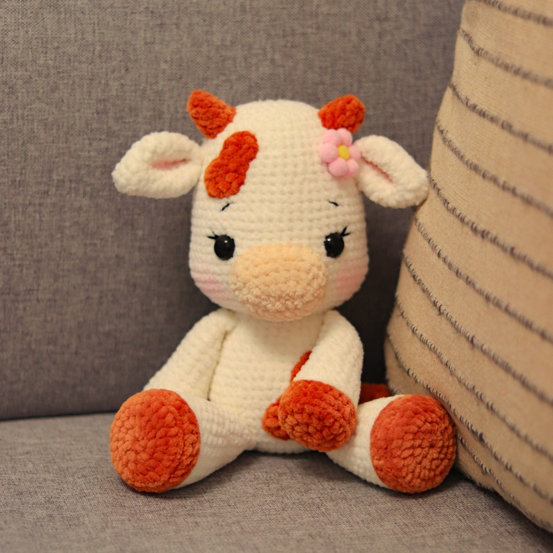 Peut inclure: Une vache en peluche crochet&eacute;e avec une fourrure blanche, des taches orange et une fleur rose sur la t&ecirc;te. La vache a des yeux noirs et un nez rose.