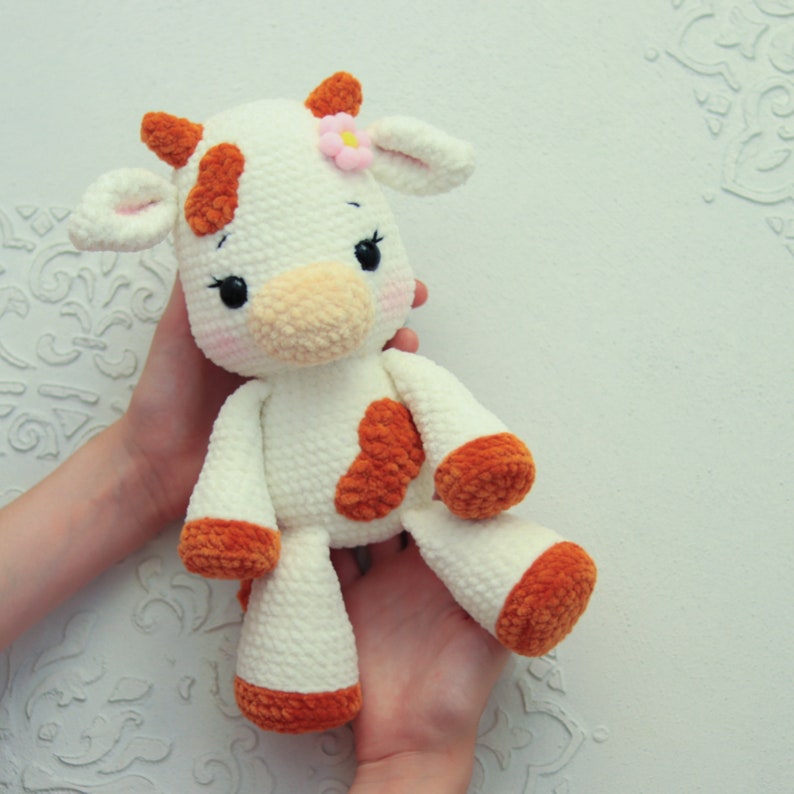Peut inclure: Un jouet en peluche de vache crochet&eacute; blanc avec des taches brunes et une fleur rose sur la t&ecirc;te. Le jouet a un patch en forme de c&oelig;ur sur la poitrine et est tenu par les mains d'une personne.