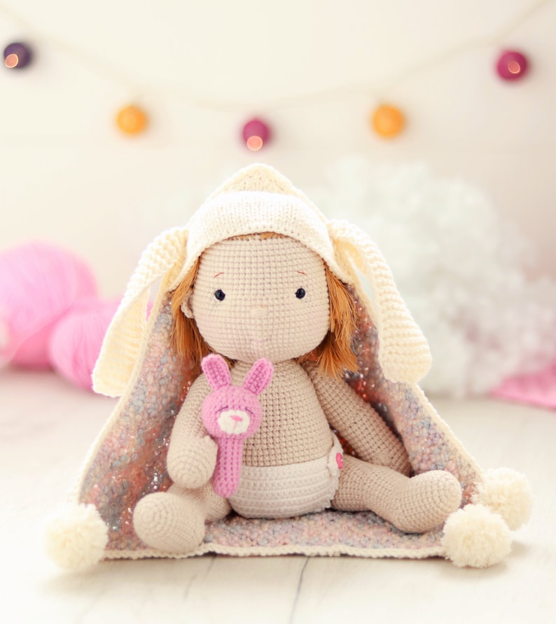 Peut inclure: Une poup&eacute;e en crochet portant une couverture &agrave; capuche blanche avec des oreilles de lapin. La poup&eacute;e tient un jouet en crochet rose en forme de lapin. La poup&eacute;e est assise sur une couverture en crochet rose et blanche.