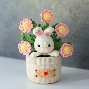 May include: A crocheted bunny peeks out from a beige pot filled with pink and yellow flowers and green leaves. The pot has a small banner with a red heart. The bunny has pink accents on its ears and nose.