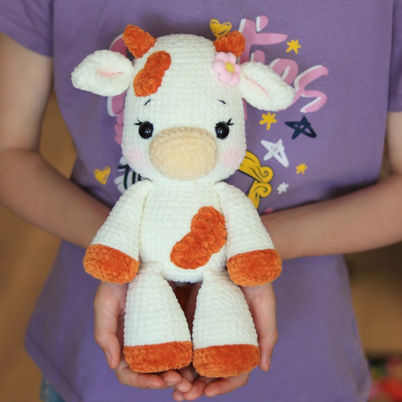 Peut inclure: Un jouet en peluche de vache blanche en crochet avec des taches brunes et une fleur rose sur la t&ecirc;te. Le jouet est tenu par les mains d'une personne.