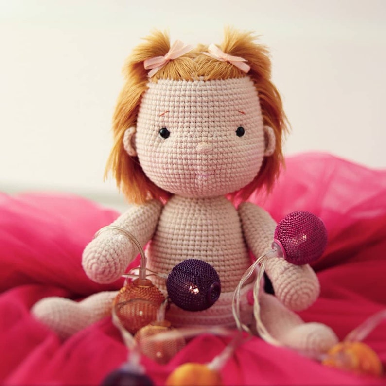Peut inclure: Une poup&eacute;e en crochet aux cheveux blonds et aux n&oelig;uds roses est assise sur un fond de tissu rose. La poup&eacute;e tient une guirlande lumineuse avec des ampoules color&eacute;es.