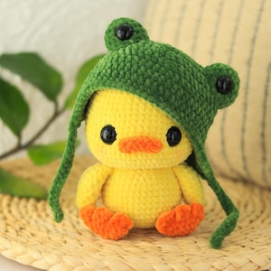 Könnte beinhalten: Eine handgefertigte gelbe Häkelente mit einem grünen Froschhut. Die Ente hat orangefarbene Füße und einen passenden Schnabel. Die Augen sind schwarz. Das Plüschtier sitzt auf einer gewebten Matte.