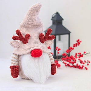 Könnte beinhalten: Ein gehäkelter Gnom mit einem rosa Hut und roten Geweih. Der Gnom hat einen weißen Bart und trägt einen gestreiften Pullover. Der Gnom sitzt auf einer weißen Oberfläche.