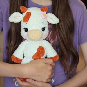 Peut inclure: Un jouet en peluche de vache blanche en crochet avec des taches brunes et une fleur rose. Le jouet a des yeux noirs et un nez brun. Le jouet est tenu par une personne.