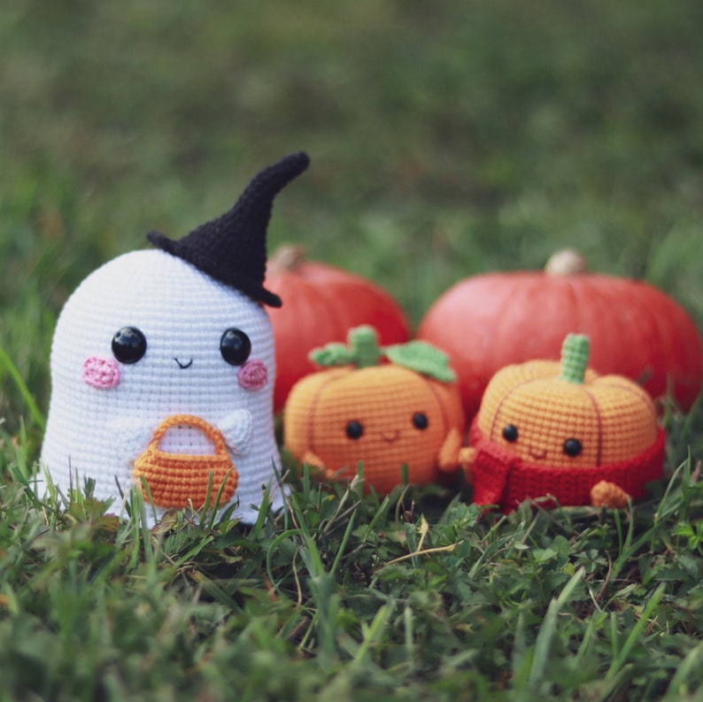 May include: A white crocheted ghost wearing a black witch hat holds an orange crocheted trick-or-treat bag. The ghost is surrounded by three crocheted pumpkins, two orange and one yellow, all with smiling faces.