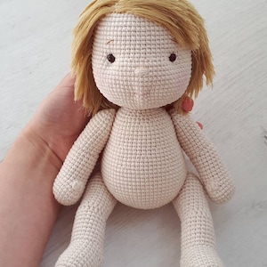 Peut inclure: Une poup&eacute;e en crochet avec des cheveux en laine blonde, assise les jambes crois&eacute;es. La poup&eacute;e a un corps beige et des yeux bruns.