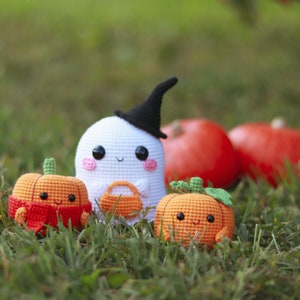 May include: A white crocheted ghost with a black witch hat, holding a small orange crocheted pumpkin. Two orange crocheted pumpkins with smiling faces are in the foreground.