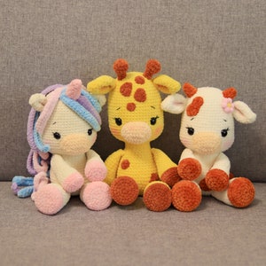 Op de afbeelding: Drie gehaakte knuffels: een eenhoorn met een roze en blauwe manen, een giraffe met gele vacht en bruine vlekken en een koe met witte vacht en bruine vlekken. De knuffels zitten op een grijze bank.