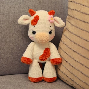 Peut inclure: Un jouet en peluche de vache crochet&eacute; avec une fourrure blanche, des taches orange et une fleur rose sur la t&ecirc;te.