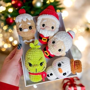 Puede incluir: Cinco adornos navideños de crochet en una bandeja blanca. Los adornos son un hombre de jengibre, Santa Claus, una mujer con cabello blanco, el Grinch y un muñeco de nieve.