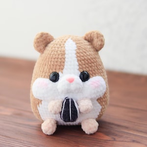 May include: A handmade crochet hamster plushie in shades of brown and white, with a pink nose and rosy cheeks. The plushie is holding a black and white sunflower seed. The hamster has large black eyes and small, rounded ears.
